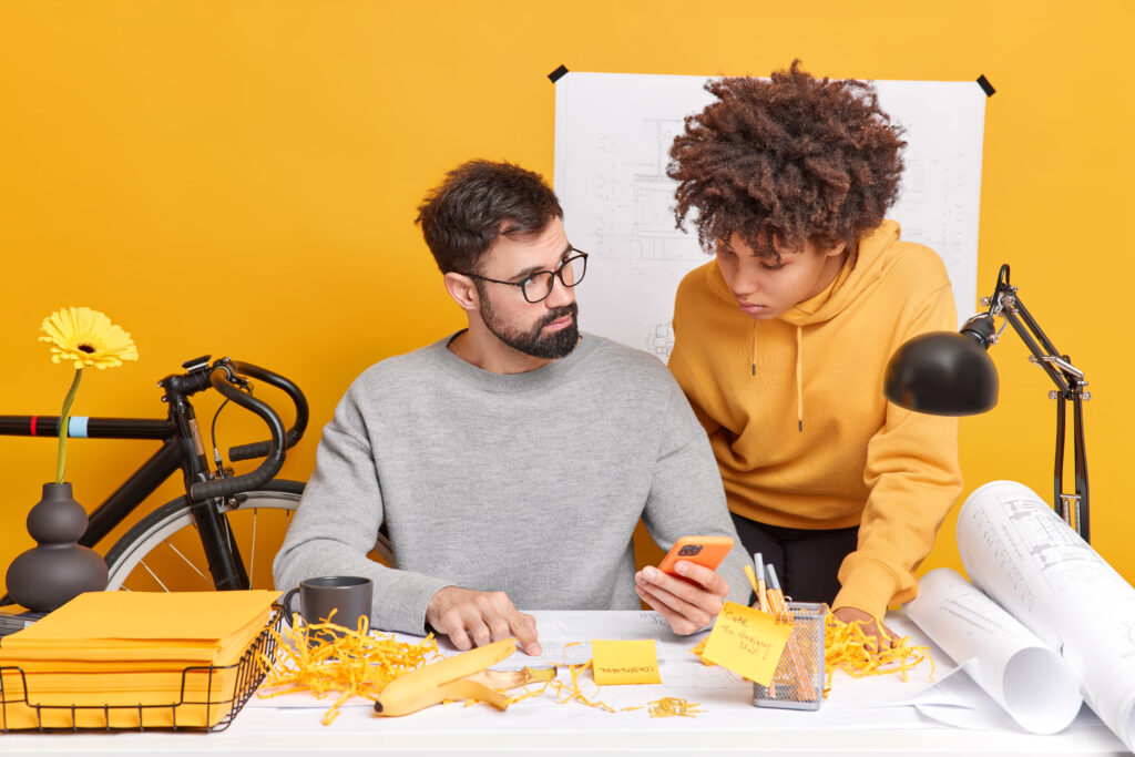 mixed race woman and man coworkers try to find solution of work check information in internet via mobile phone study papers pose in coworking space enjoy cooperation isolated over yellow wall